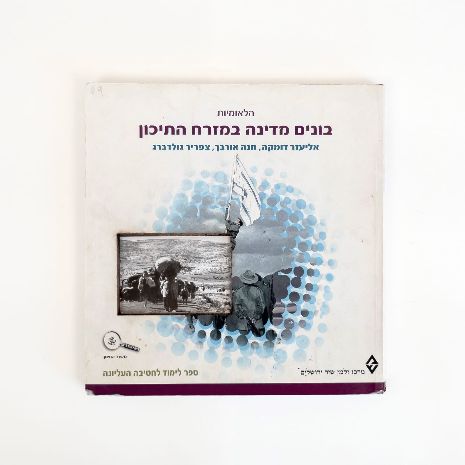Photograph of a mostly white Israeli history textbook against a white background. The Hebrew title reads in dark purple, The Nationalism, and then, Building a State in the Middle East. Centered on the cover is the photograph of the raising of the ink flag around the time of establishment of the state of Israel. A rectangle is cut into a part of the image and reveals a photograph from deeper in the book of the 1948 Palestinian Nakba: people walking on a dirt road past a broken car carrying belongings.