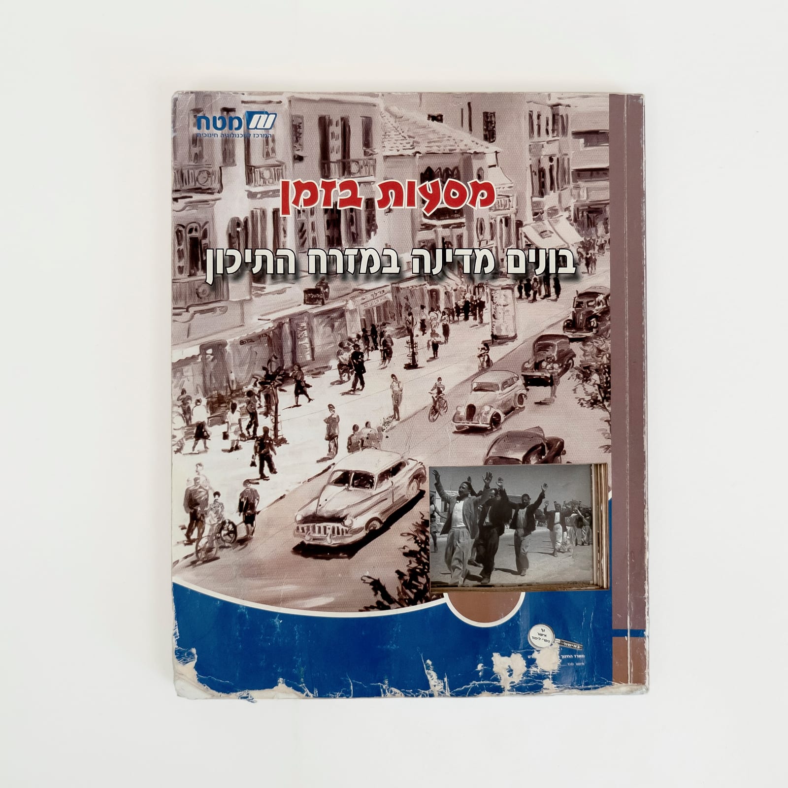 Photograph of a worn Israeli history textbook against a white background.A rectangular cut in the bottom right area of the cover to reveal a photograph of displaced Palestinians walking with their arms raised during the 1948 Nakba. The Hebrew title reads in red and white, Time Travels: Building a State in the Middle East. The majority of the cover is a realistic sepia drawing of a street with cars and people walking. The bottom of the cover is blue.