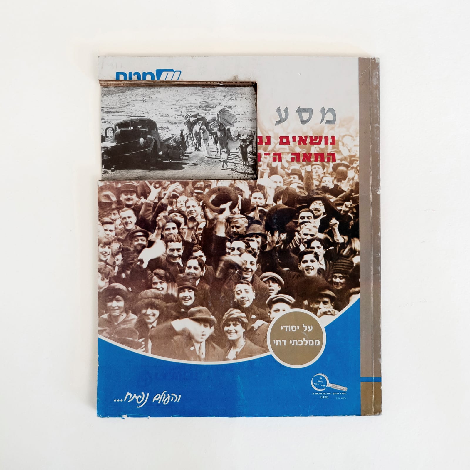 Photograph of an Israeli history textbook against a white background. A rectangular cut at top left edge of the book reveals a photograph from inside the book of the 1948 Palestinian Nakba: people walking on a dirt road carrying belongings, with a broken down car on the side of the road. The cut leaves parts of the Hebrew title legible: Journey in gray, and then, Topics, the century. The cover features a full bleed sepia photograph of a dense crowd in celebration. The bottom of the cover is blue.