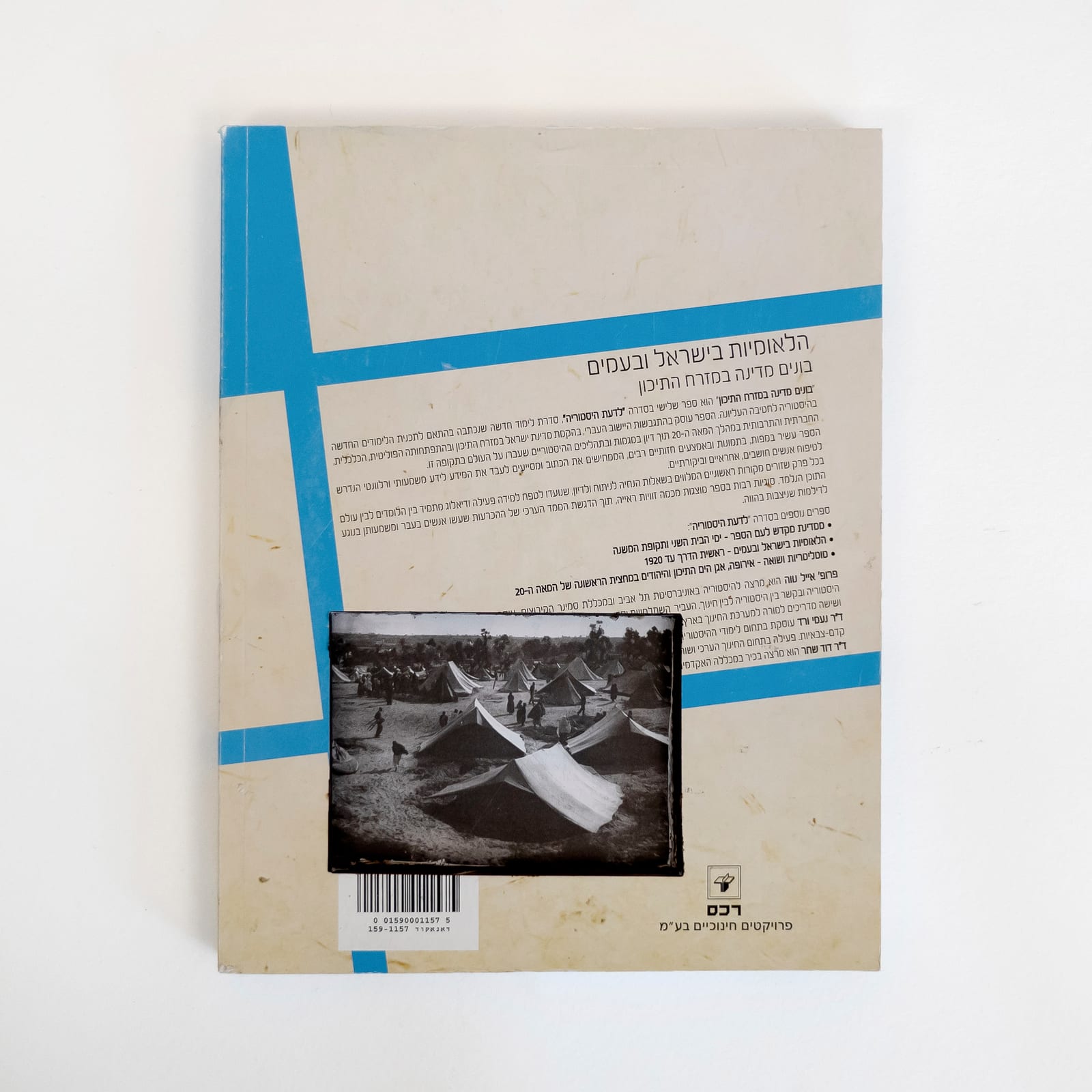 Photograph of the back of an Israeli history textbook against a white background. A block of Hebrew text is set against a tan color between blue rules. A rectangle has been cut down through the cover to reveal a photograph of the 1948 Palestinian Nakba from deeper in the book showing a tent encampment.